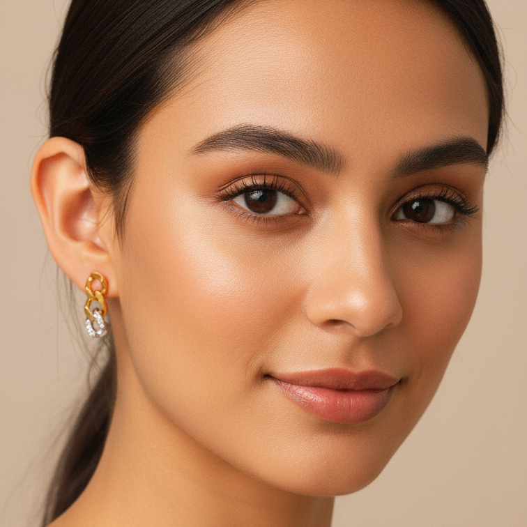 A close-up portrait of a woman with dark hair pulled back, wearing a Gold & Silver Pavé Chain Link Earrings. The background is a solid warm beige tone.