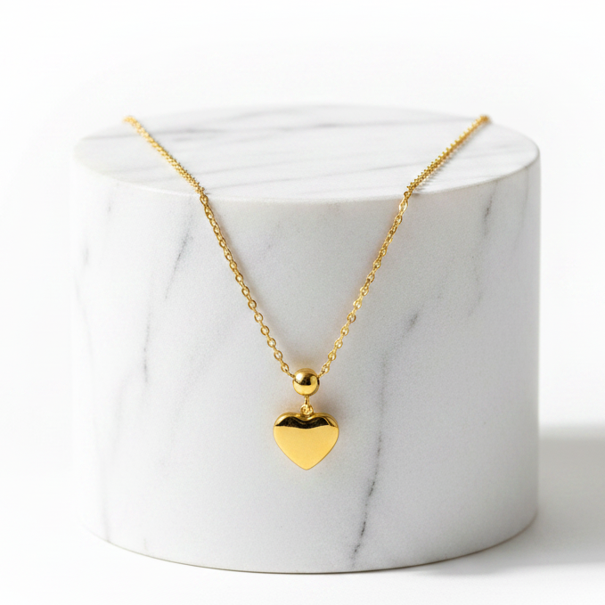A gold heart pendant necklace on a delicate gold chain, displayed on a white marble cylinder pedestal against a clean white background.