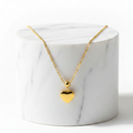 A gold heart pendant necklace on a delicate gold chain, displayed on a white marble cylinder pedestal against a clean white background.