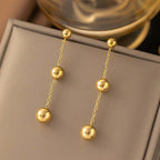 Gold Ball Tassel Earrings with three polished gold spheres on delicate chain, displayed on beige background