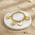 Gold Coastal Charm Bracelet on a white stone with seashells and sand in the background