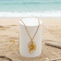 Gold Conch Shell Pendant Necklace displayed on a white pedestal against a beach background.