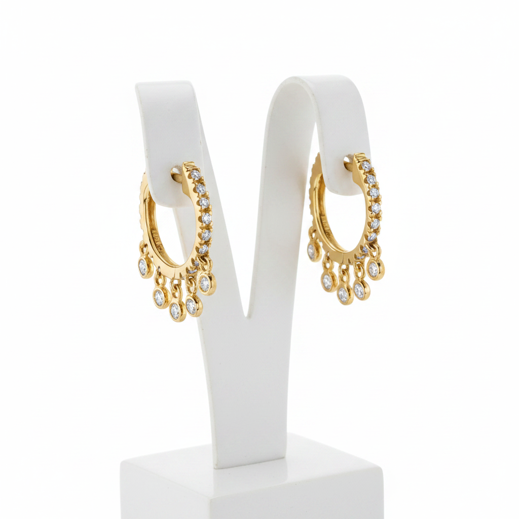 A pair of gold huggie hoop earrings adorned with clear pavé stones and dangling teardrop accents, displayed on a white V-shaped jewelry stand against a white background.