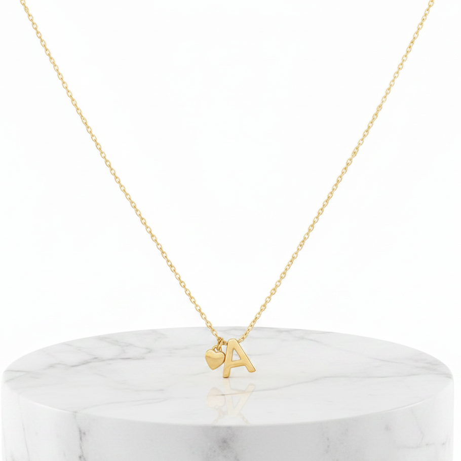 A delicate gold chain necklace with a small initial pendant featuring the letter 'A' and a small heart charm, displayed on a white marble cylinder pedestal against a white background.