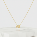 A delicate gold chain necklace with a cutout letter B pendant and a small heart charm, displayed on a white marble block against a light cream background.