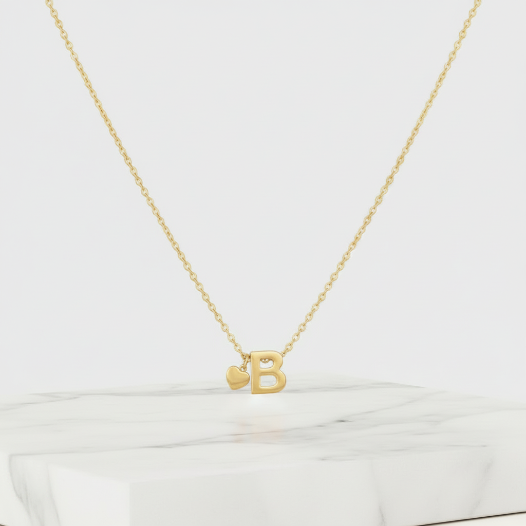 A delicate gold chain necklace with a cutout letter B pendant and a small heart charm, displayed on a white marble block against a light cream background.