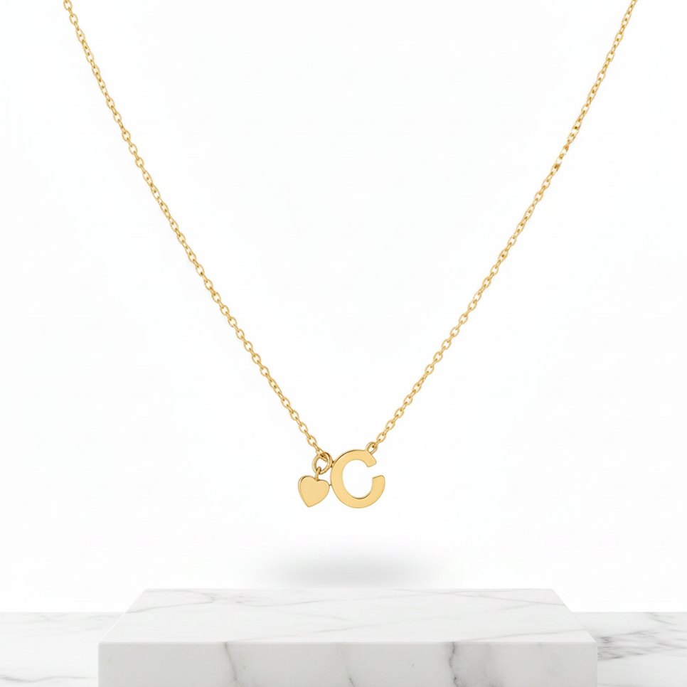 A delicate gold chain necklace featuring a small letter 'C' pendant and a small heart charm, displayed on a white marble block against a clean white background.