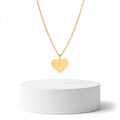 A Gold Heart Heartbeat Necklace engraved with an EKG line, resting on a white surface with a textured stone prop in the corner.