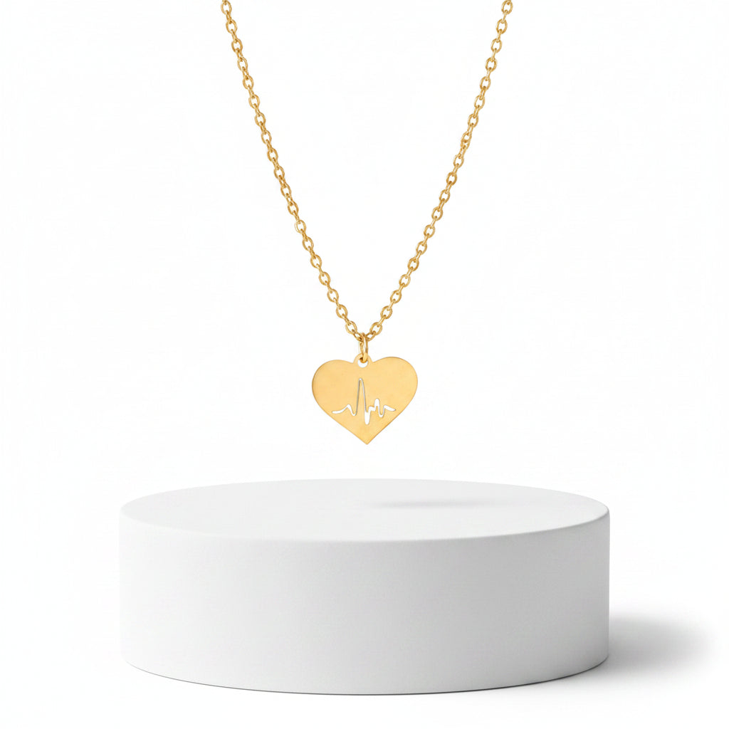 A Gold Heart Heartbeat Necklace engraved with an EKG line, resting on a white surface with a textured stone prop in the corner.