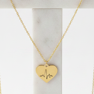 A gold Gold Heart Heartbeat Necklace with an EKG line engraving, displayed on a white marble jewellery stand against a clean white background.