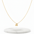 A delicate gold chain necklace with a small pendant featuring a heart and a vertical bar, displayed on a round white marble stand against a clean white background.