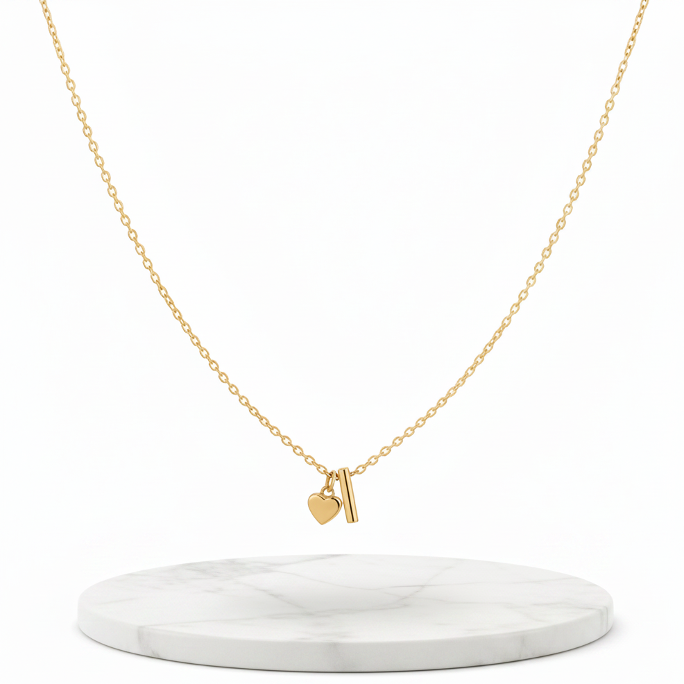 A delicate gold chain necklace with a small pendant featuring a heart and a vertical bar, displayed on a round white marble stand against a clean white background.