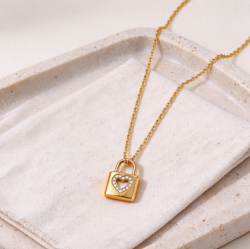 A Gold Heart Padlock Necklace featuring a heart-shaped crystal center, displayed on a textured cream fabric over a beige stone tray.