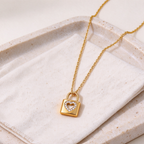 A Gold Heart Padlock Necklace featuring a heart-shaped crystal center, displayed on a textured cream fabric over a beige stone tray.