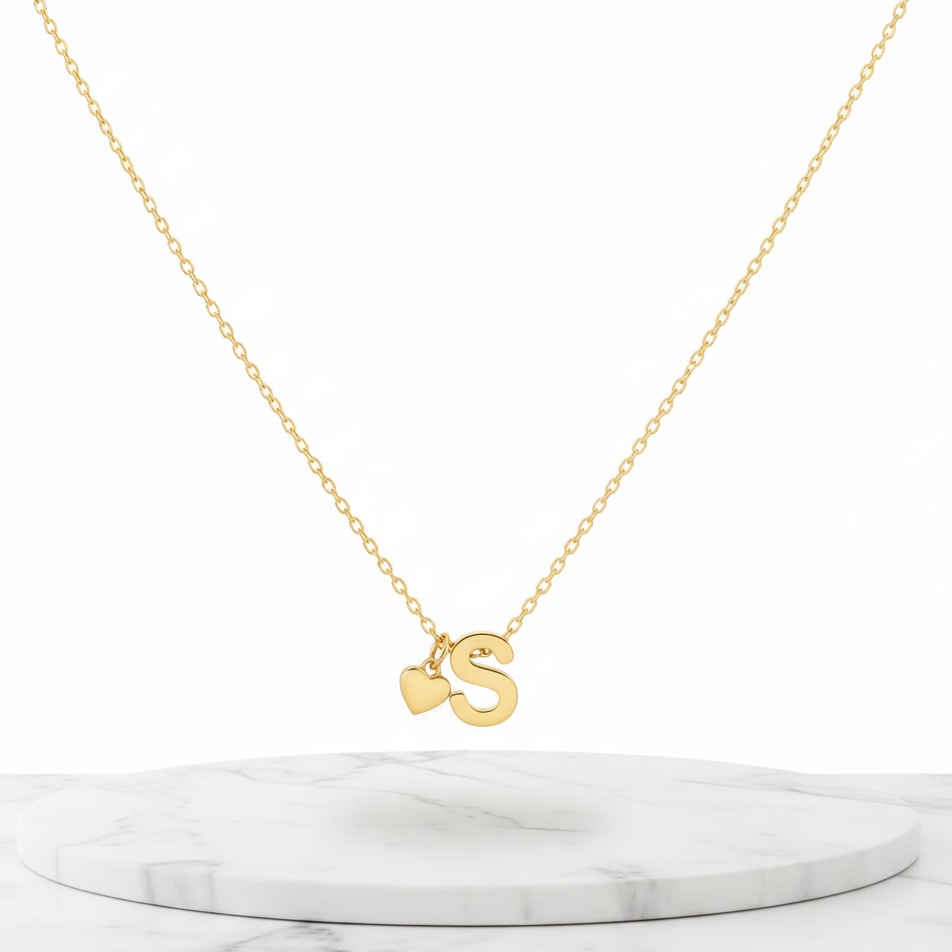 Gold Heart & S Initial Pendant Necklace displayed on a white marble pedestal against a white background.