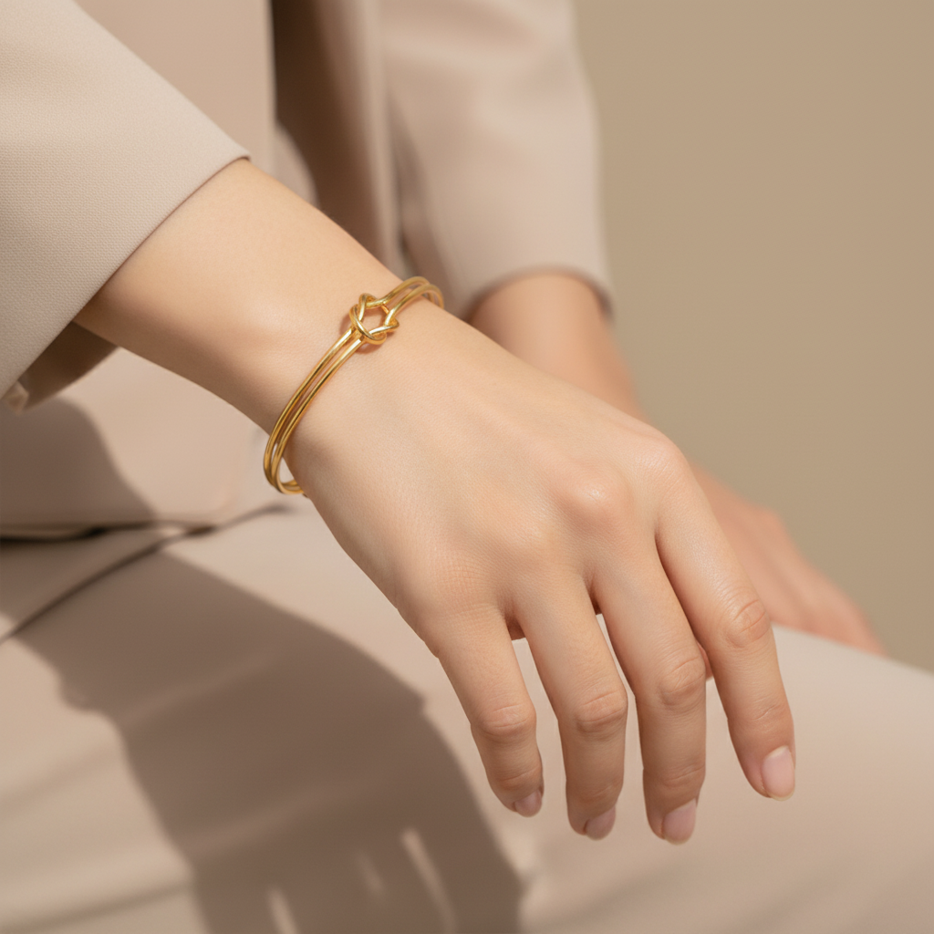 A close-up of a person's hand and wrist wearing a delicate gold bangle bracelet with a geometric openwork connector, set against a soft beige background.