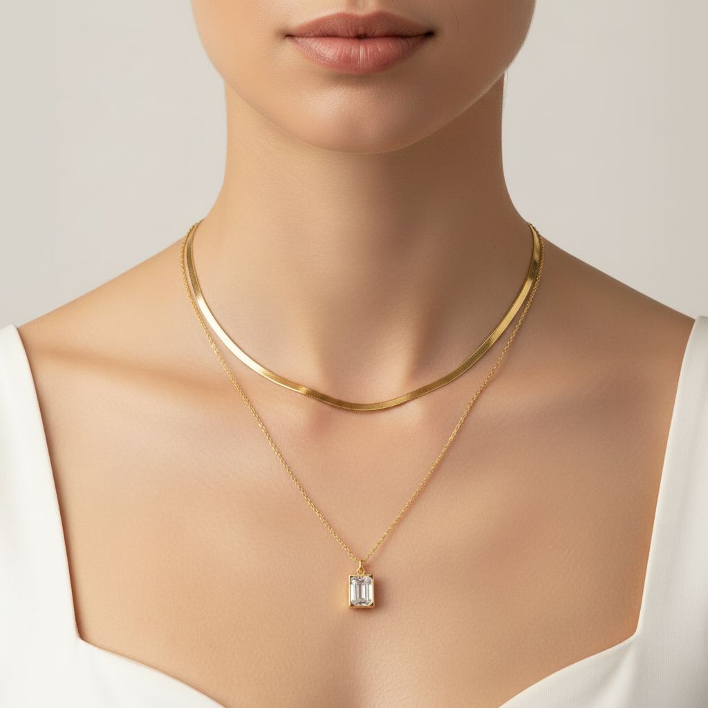 A close-up of a model's neck and chest wearing a layered gold necklace set, featuring a thick curved collar choker and a delicate chain with an emerald-cut clear gemstone pendant.