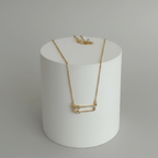 Gold Safety Pin Pendant Necklace on a white cylindrical stand against a gray background