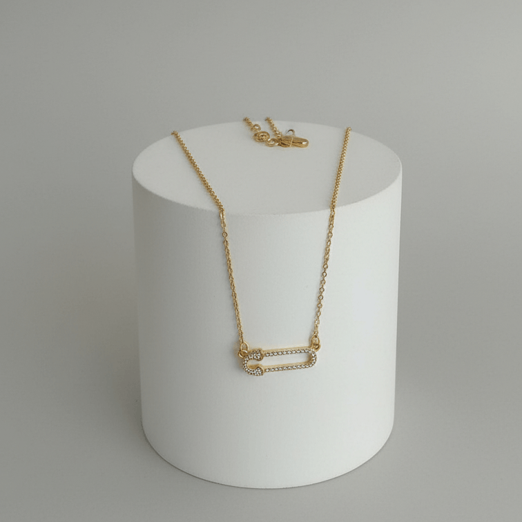 Gold Safety Pin Pendant Necklace on a white cylindrical stand against a gray background