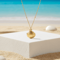 Gold Scallop Shell Pendant Necklace with a shell pendant on a white platform by the beach
