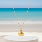 Gold Scallop Shell Pendant Necklace with a shell pendant on a white pedestal against a beach background