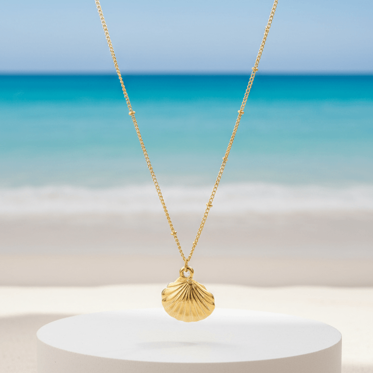 Gold Scallop Shell Pendant Necklace with a shell pendant on a white pedestal against a beach background