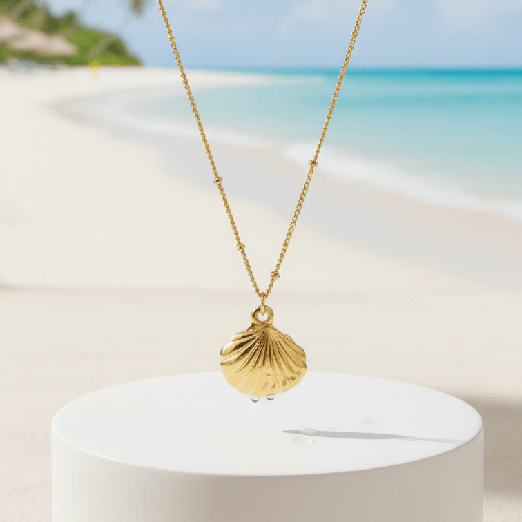 Gold Scallop Shell Pendant Necklace with a shell pendant on a white pedestal against a beach background