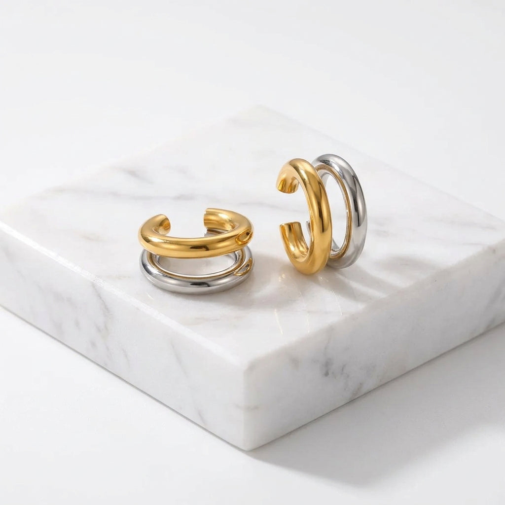 A pair of Gold & Silver Double Layered Ear Cuff with a polished gold top band and a silver-toned lower band, displayed on a textured beige surface.