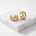A pair of Gold & Silver Double Layered Ear Cuff with a polished gold top band and a silver-toned lower band, displayed on a textured beige surface.