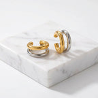 A pair of Gold & Silver Double Layered Ear Cuff with a polished gold top band and a silver-toned lower band, displayed on a textured beige surface.