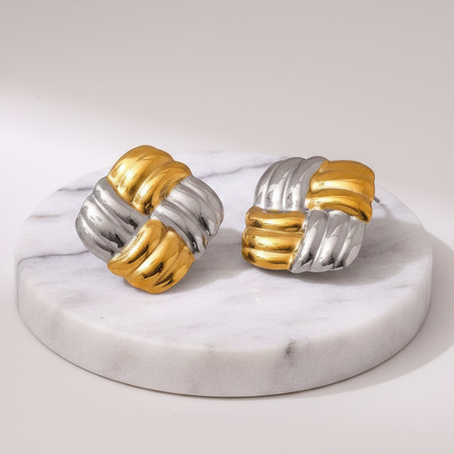A pair of two-tone twisted knot earrings in polished gold and silver, displayed on a round white marble stand against a light gray background.