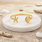 Gold Starfish & Shell Bangle Bracelet on a white stone with a beach background