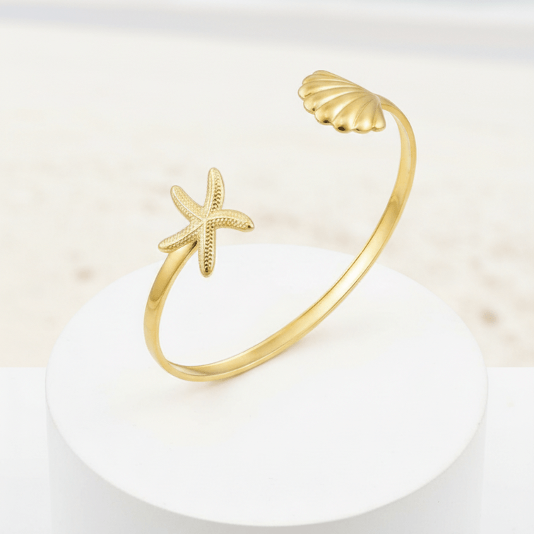 Gold bracelet with starfish and shell designs on a white stand with a beach background