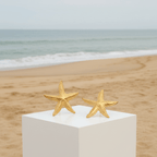 Gold Starfish Stud Earrings on a white pedestal with a beach background