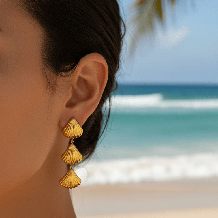 Close-up of a person wearing Gold Tiered Seashell Earrings with a beach in the background
