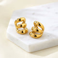 Gold Triple Band Ear Cuff displayed on a light beige surface, with one earring resting on a textured stone block and the other placed beside it.