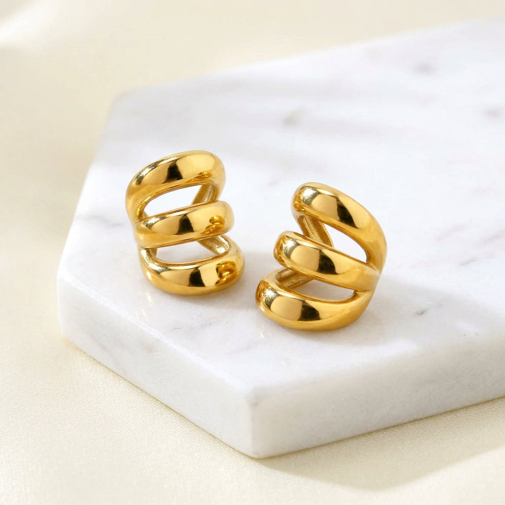 Gold Triple Band Ear Cuff displayed on a light beige surface, with one earring resting on a textured stone block and the other placed beside it.