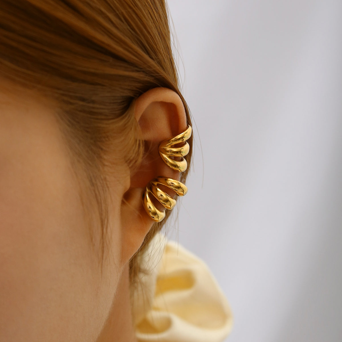 A close-up of a model's ear wearing a Gold Triple Band Ear Cuff against a soft, light gray background.
