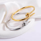 Bread Nail Bangle Bracelet in gold and silver stainless steel on a soft satin surface.