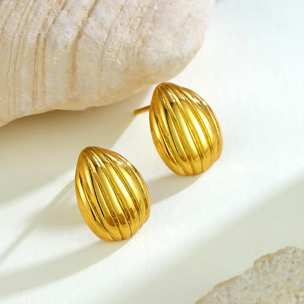 Gold Ribbed Teardrop Stud Earrings on a light beige surface with a shell in the background
