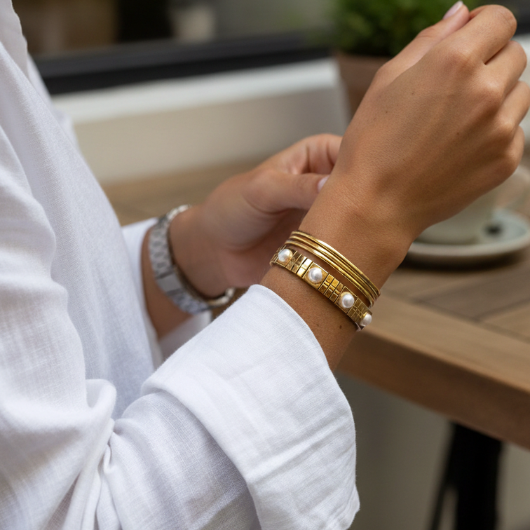 A close-up lifestyle shot of a person wearing a stack of gold bangles, including one with white pearls, on their wrist. The person is wearing a white linen shirt and sitting at a wooden table in a cafe setting.