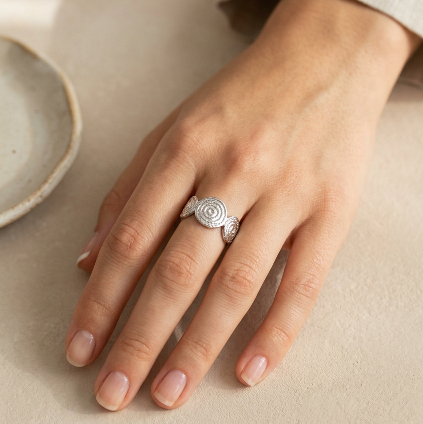 A close-up of a hand wearing a Silver Spiral Adjustable Ring, set against a warm beige background with a ceramic dish partially visible on the left.