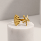 Gold shell and starfish ring on a white cylindrical stand with a neutral background
