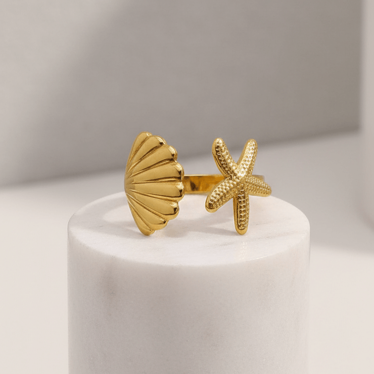 Gold shell and starfish ring on a white cylindrical stand with a neutral background