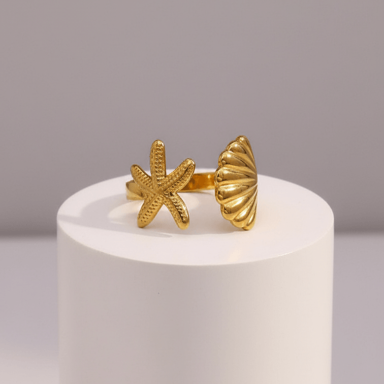 Gold starfish and shell ring on a white cylindrical stand with a gray background