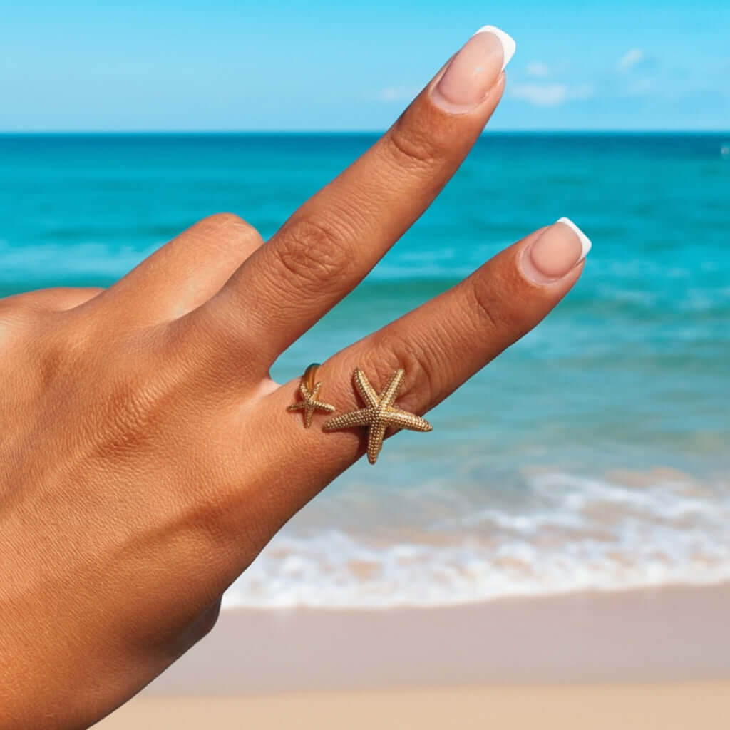 Hand wearing a Twin Starfish Open Gold Ring with a beach background