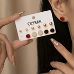 A close-up of a person wearing a pair of Waterproof Gold Circle Stud Earrings Set with red and black textured centers, while another hand holds a white display card showing additional matching earrings. The card is branded 'CEYRAH'.