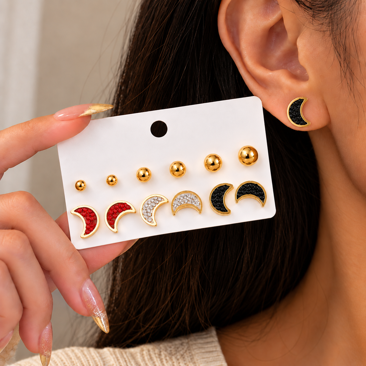 A close-up of a person wearing a gold crescent moon stud earring, while holding a white display card showing Waterproof Gold Moon Stud Earrings Set in red, silver, and black enamel.
