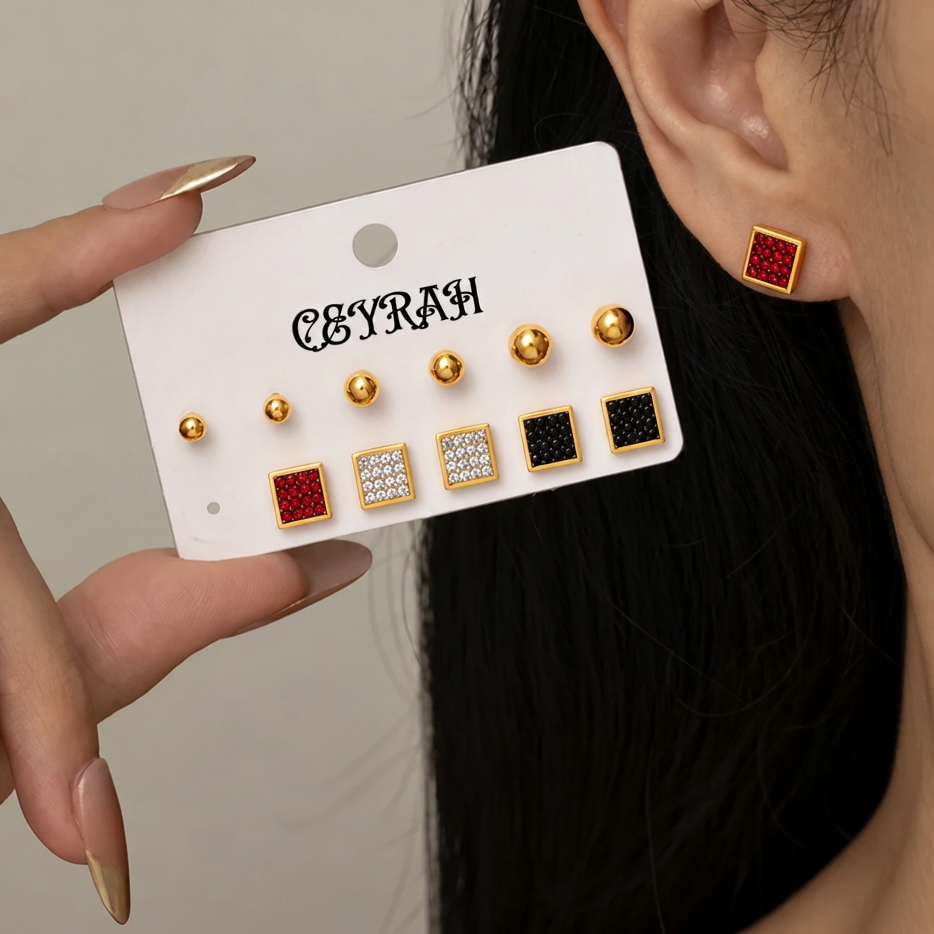 A close-up of a person wearing a pair of Waterproof Gold Square Stud Earrings Set with red and black textured centers, while another hand holds a white display card showing additional matching earrings. The card is branded 'CEYRAH'.