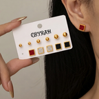 A close-up of a person wearing a pair of Waterproof Gold Square Stud Earrings Set with red and black textured centers, while another hand holds a white display card showing additional matching earrings. The card is branded 'CEYRAH'.